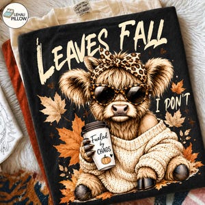 May include: A black t-shirt featuring a graphic of a fluffy highland cow wearing a leopard print headband and sunglasses. The cow wears a beige sweater, holds a coffee cup labeled "Fueled by CHAOS" with a pumpkin icon, and is surrounded by autumn leaves with the text "LEAVES FALL I DON'T".