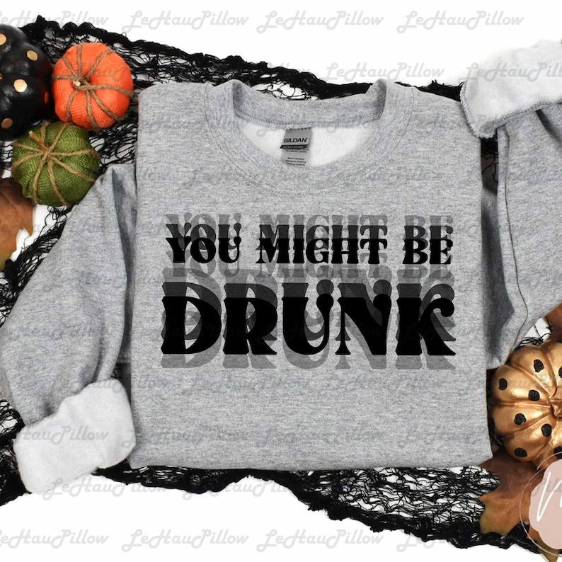 May include: A gray sweatshirt with the text "YOU MIGHT BE DRUNK" in black. The sweatshirt is surrounded by Halloween decorations, including pumpkins and a black decorative net.