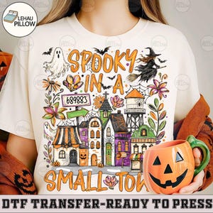 May include: A cream-colored t-shirt featuring a colorful Halloween-themed graphic with the text "SPOOKY IN A SMALL TOWN". The design includes a ghost, a witch on a broom, a water tower, various buildings, flowers, and a sign labeled "689883".