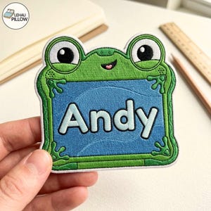 May include: A green frog-shaped embroidered patch featuring a blue rectangular centre with the name Andy stitched in light blue letters. The frog has large white eyes with black pupils and a small smiling mouth. The patch has a white border.