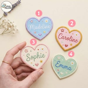 Könnte beinhalten: Vier herzförmige Stickaufnäher mit gewelltem Rand. Jeder Aufnäher zeigt einen Namen in Schreibschrift: Madeline in Blau, Caroline in Pink, Sophie in Creme und Emma in Grün. Kleine Blumen- und Sternmotive verzieren jedes der Herzen.