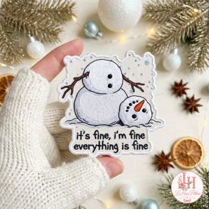 May include: A white embroidered patch featuring a snowman with a detached head resting on the ground. The snowman has brown stick arms and black eyes. The text reads: it's fine, i'm fine everything is fine. The patch has a scalloped edge.