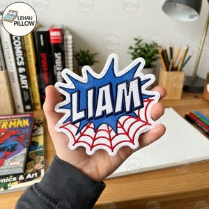 May include: A custom embroidered patch featuring the name LIAM in bold white letters with a dark blue outline, set against a blue comic book style burst shape and a red spider web design on a white background with a thick white border.