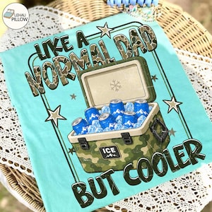 May include: A light blue t-shirt with the text "LIKE A NORMAL DAD BUT COOLER". The graphic features a camouflage cooler filled with ice and blue cans. The design includes star accents.
