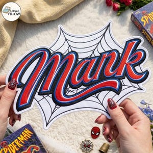 May include: A white embroidered patch featuring a black spiderweb background and the name Mark in red lettering with blue and black outlines. The patch has a distinct comic book style font and is held against a textured white surface.
