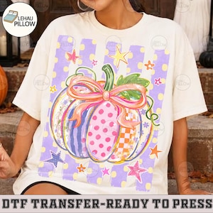 May include: A cream-colored short-sleeve t-shirt featuring a large graphic of a pumpkin decorated with pink polka dots, orange checkered patterns, and a pink bow. The design includes purple vertical stripes and various colorful stars in the background.