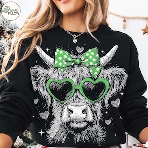 May include: Black sweatshirt featuring a detailed illustration of a Highland cow with heart-shaped sunglasses and a green bow. The cow's fur is rendered in intricate detail, with a whimsical design. The design includes small hearts and stars.