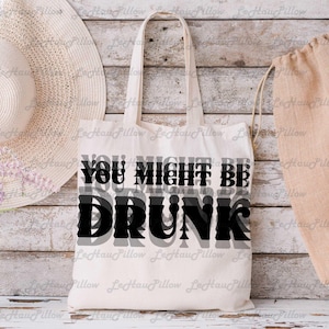 May include: Cream-colored tote bag with the text "YOU MIGHT BE DRUNK" in black and gray. The bag has two handles and is made of a canvas-like material. A straw hat and a drawstring bag are in the background.