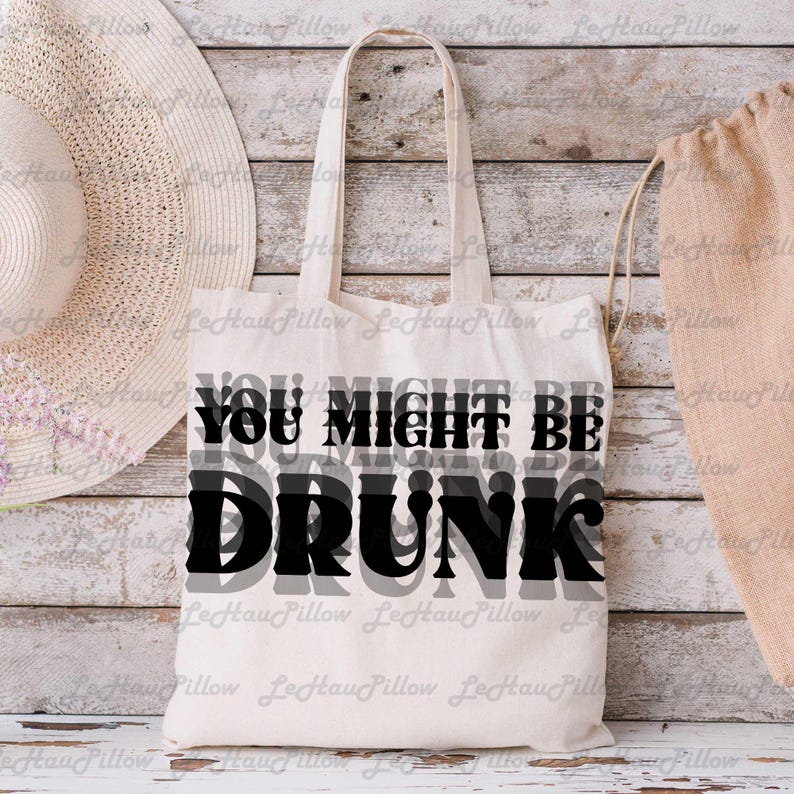 May include: Cream-colored tote bag with the text "YOU MIGHT BE DRUNK" in black and gray. The bag has two handles and is made of a canvas-like material. A straw hat and a drawstring bag are in the background.