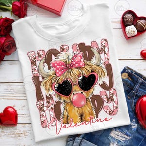 May include: White t-shirt with a graphic of a Highland cow wearing heart-shaped sunglasses and a pink bow. The text "Happy Valentine" is displayed in pink and brown. A bubblegum bubble is visible.