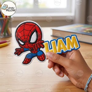 May include: An embroidered fabric patch featuring a cartoon Spider-Man character in red and blue with a web pattern, positioned next to the name LIAM in yellow block letters with a blue border, all outlined in white thread on a fabric backing.
