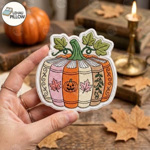 May include: An embroidered patch shaped like a pumpkin made of stacked books. The books feature autumn colors including orange, cream, and pink, with details like a jack-o'-lantern, maple leaves, and vines. A green stem and leaves sit on top.