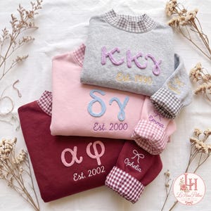 Pode incluir: Três camisolas em cinzento, rosa e bordeaux, cada uma com letras gregas bordadas, um ano de fundação e gola e punhos com padrão xadrez vichy. As mangas são personalizadas com nomes bordados e ícones de laços.