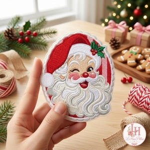 May include: An embroidered patch featuring a winking Santa Claus with a white beard, red hat, and green holly leaves with red berries. The circular patch has a scalloped white border and is held up against a festive holiday background.