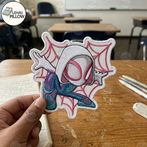 May include: An embroidered patch featuring a stylized character wearing a white hood with pink accents, a blue and purple suit with a spider web pattern, and a pink spider web background. The character is in a dynamic pose with arms outstretched.
