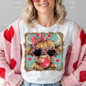 Peut inclure: T-shirt blanc avec une vache des Highlands de dessin anim&eacute; portant des lunettes de soleil en forme de c&oelig;ur et une bulle de chewing-gum. La vache a un n&oelig;ud rouge et est entour&eacute;e de c&oelig;urs. Th&egrave;me de la Saint-Valentin.