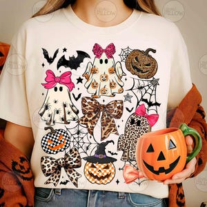 May include: A cream-colored t-shirt featuring a graphic print of three ghosts, pumpkins, leopard print bows, spiderwebs, and bats. One ghost wears a pink bow, another has a leopard print pattern, and a third has a pumpkin print. An orange pumpkin mug is held.