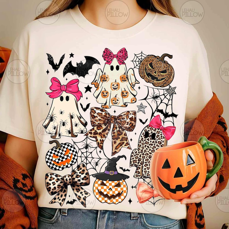 May include: A cream-colored t-shirt featuring a graphic print of three ghosts, pumpkins, leopard print bows, spiderwebs, and bats. One ghost wears a pink bow, another has a leopard print pattern, and a third has a pumpkin print. An orange pumpkin mug is held.