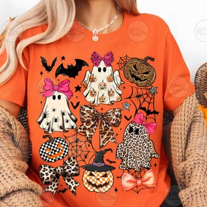 May include: An orange t-shirt featuring a graphic print of three ghosts, two pumpkins, and several bows. The ghosts have patterns including spiders, small pumpkins, and leopard print. The pumpkins feature checkered and leopard patterns.