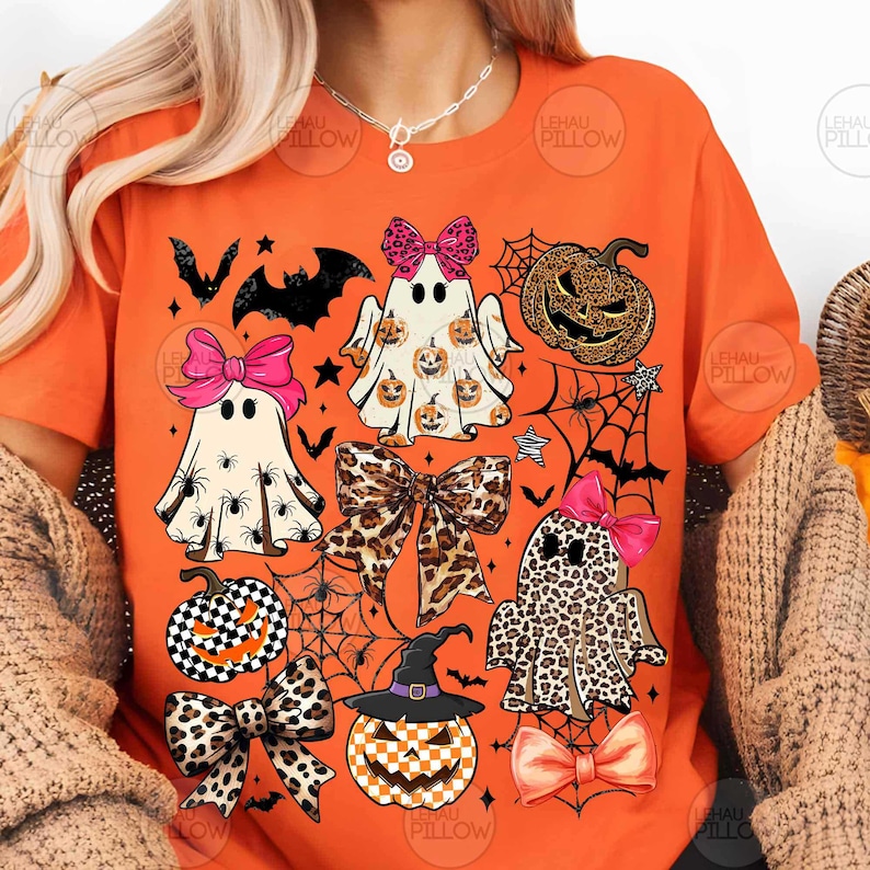 May include: An orange t-shirt featuring a graphic print of three ghosts, two pumpkins, and several bows. The ghosts have patterns including spiders, small pumpkins, and leopard print. The pumpkins feature checkered and leopard patterns.