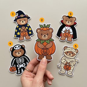Pode incluir: Cinco emblemas bordados com ursinhos de peluche castanhos em trajes de Halloween, cada um segurando um pequeno balde de abóbora laranja. Os ursos estão vestidos de bruxa, abóbora, vampiro, esqueleto e múmia, numerados de 1 a 5.