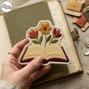 May include: An embroidered patch featuring an open book with cream-coloured pages and a dark red cover. Three flowers emerge from the top of the book: a central yellow flower with a red centre, flanked by two red tulip-like flowers on green stems.