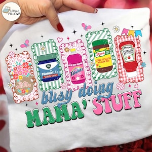 Könnte beinhalten: Weißes T-Shirt mit einem farbenfrohen Grafikdesign. Das Design zeigt verschiedene Produktillustrationen, darunter ein Glas MamaRub, eine Flasche Fabulosa und eine Flasche Mama Tomato Ketchup. Der Text "busy doing MAMA' STUFF" wird ebenfalls angezeigt.