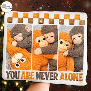 Könnte beinhalten: Ein weißes Kissen mit einem Design, das vier Plüschaffen in Orange und Grau zeigt, die sich umarmen. Der Text "YOU ARE NEVER ALONE" ist unten in Braun gedruckt. Der obere Rand hat ein Schachbrettmuster.