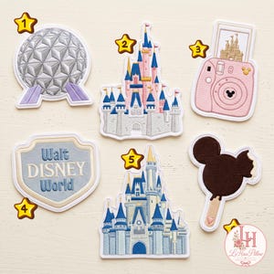Peut inclure: Un ensemble de six écussons brodés représentant une sphère géodésique argentée, un château rose et bleu, un appareil photo instantané rose avec une photo de château, un écusson avec le texte Walt Disney World, un château bleu et blanc et une glace.