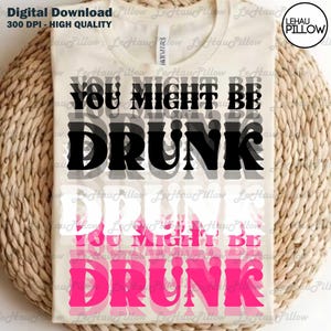 May include: Cream-colored t-shirt with the text "YOU MIGHT BE DRUNK" in black and pink. The design is layered, creating a shadow effect. The shirt is displayed on a woven surface.
