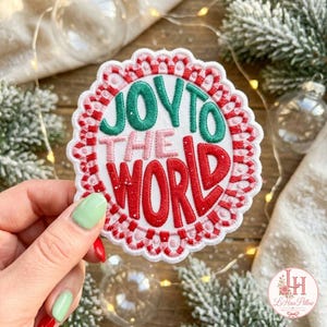 Könnte beinhalten: Ein runder Stickaufnäher mit gewelltem Rand und einem rot-weißen Zuckerstangenmuster. Die weiße Mitte zeigt den Text JOY TO THE WORLD in grünen, rosa und roten gestickten Buchstaben mit einem dekorativen Randdesign.