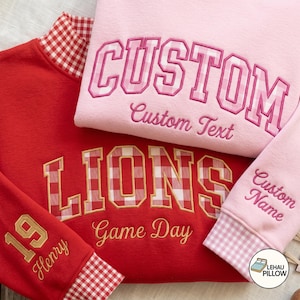 May include: A red sweatshirt with a white and red gingham applique reading LIONS and Game Day, plus a pink sweatshirt with a pink gingham applique reading CUSTOM and Custom Text. Both feature embroidered names and numbers on the sleeves.