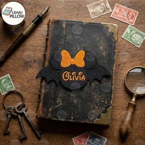 May include: A black embroidered patch featuring a circular center with the name Olivia in orange, flanked by bat wings and topped with an orange bow. The patch is centered on a weathered, dark-bound vintage book with metal corner accents.
