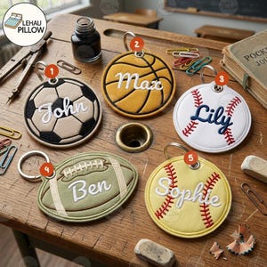 May include: Five circular and oval keychains featuring embroidered sports designs and names. The keychains include a soccer ball with John, a basketball with Max, a baseball with Lily, a football with Ben, and a softball with Sophie.