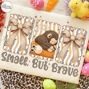May include: Cream-colored pillow with a whimsical design featuring two bows and a sleeping monkey illustration. The text "Small, But Brave" is printed below the illustrations. The pillow has a scalloped border and a striped background.