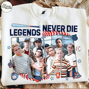 Puede incluir: Sudadera blanca con un diseño de béisbol. El diseño incluye un bate de béisbol, una pelota de béisbol, estrellas y un guante de béisbol, con el texto "LEGENDS NEVER DIE". La sudadera es de cuello redondo.