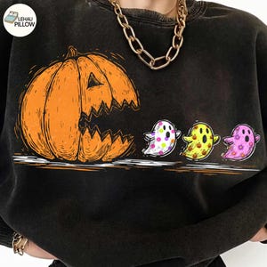 May include: A black crewneck sweatshirt featuring a large orange jack-o'-lantern graphic with a wide, jagged mouth. To the right of the pumpkin are three small ghosts with colorful patterns, including polka dots, mini pumpkins, and flowers.