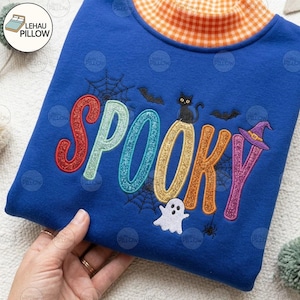 Può includere: Una felpa blu reale con collo alto a quadri arancioni e bianchi. La parte anteriore mostra la parola SPOOKY in lettere glitterate multicolori, accompagnata da pipistrelli neri ricamati, un gatto nero, un fantasma bianco e ragnatele.