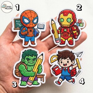 Może przedstawiać: Cztery haftowane naszywki z postaciami z kreskówek: Spider-Man z tablicą i książkami, Iron Man trzymający ołówek przy klockach z literami, Hulk z dużym ołówkiem oraz uczeń z czerwoną książką, kątomierzem i linijką.