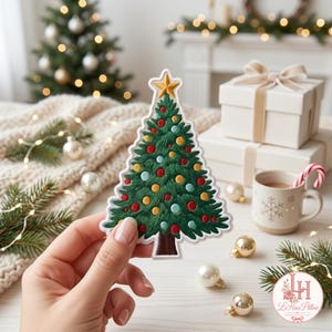 Può includere: Una toppa a forma di albero di Natale con ricamo verde, decorata con ornamenti circolari rossi, gialli e azzurri e una stella dorata in cima. La toppa presenta un bordo bianco e un tronco marrone, tenuta davanti a uno sfondo festivo.