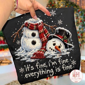 May include: A black t-shirt featuring a graphic of two snowmen, one standing and one lying down, wearing a red and green plaid scarf and a black top hat with holly. The text reads, it's fine, i'm fine, everything is fine, with white snowflakes.