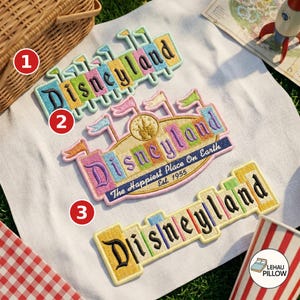 May include: Three embroidered Disneyland patches on a white fabric surface. The top patch features colorful letters and flags. The middle patch displays the Disneyland logo with the text The Happiest Place On Earth Est. 1955. The bottom patch is yellow.