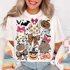 May include: A cream-colored t-shirt featuring a graphic print of three ghosts, two pumpkins, and four bows. The ghosts and pumpkins have leopard print, checkered, and spider patterns. The bows are pink, leopard print, and orange.