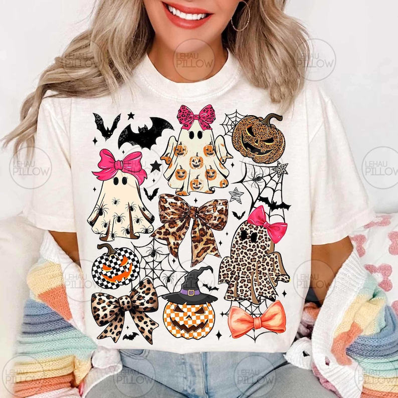 May include: A cream-colored t-shirt featuring a graphic print of three ghosts, two pumpkins, and four bows. The ghosts and pumpkins have leopard print, checkered, and spider patterns. The bows are pink, leopard print, and orange.
