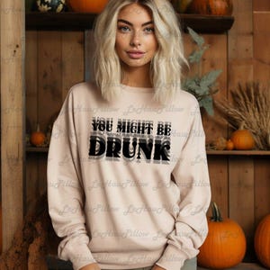 May include: A light pink sweatshirt with the text "YOU MIGHT BE DRUNK" in black and gray. The sweatshirt is a crew neck style. Pumpkins and fall decor are in the background.