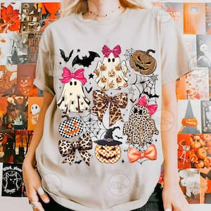 May include: A beige short-sleeve t-shirt featuring a graphic print of three ghosts, two pumpkins, and several bows. The ghosts and pumpkins are decorated with leopard print, spiderweb, and checkerboard patterns, accented by pink and orange bows.