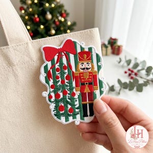 Könnte beinhalten: Ein gestickter Aufnäher mit einem Nussknacker-Soldaten neben einem grünen Weihnachtsbaum, der mit roten Kugeln und einer großen roten Schleife verziert ist, vor einem Hintergrund aus vertikalen grünen und weißen Streifen auf einer Stofftasche.
