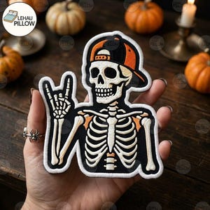 May include: An embroidered patch featuring a skeleton wearing an orange and black baseball cap, making a rock-and-roll hand gesture. The skeleton is depicted in white thread against a black background with orange accents on the shoulders and cap.
