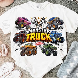 Może przedstawiać: Biała koszulka z kolorową grafiką przedstawiającą sześć monster truck&oacute;w w r&oacute;żnych kolorach, w tym r&oacute;żowym, pomarańczowym i zielonym. Tekst "MONSTER TRUCK" jest wyraźnie widoczny. Szachownice i błyskawice dopełniają projekt.
