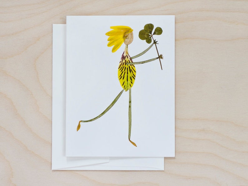 May include: A whimsical greeting card featuring a person made of pressed flowers. The person is holding a four-leaf clover and has yellow hair and a yellow and green dress.